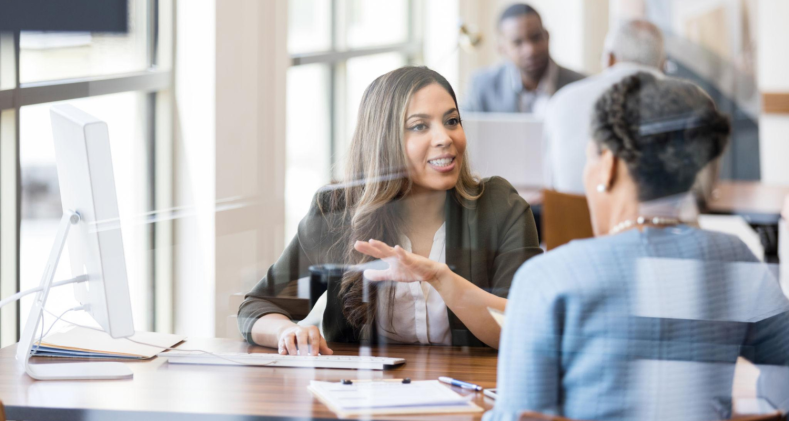Two women have a conversation at a table in a financial company's office.